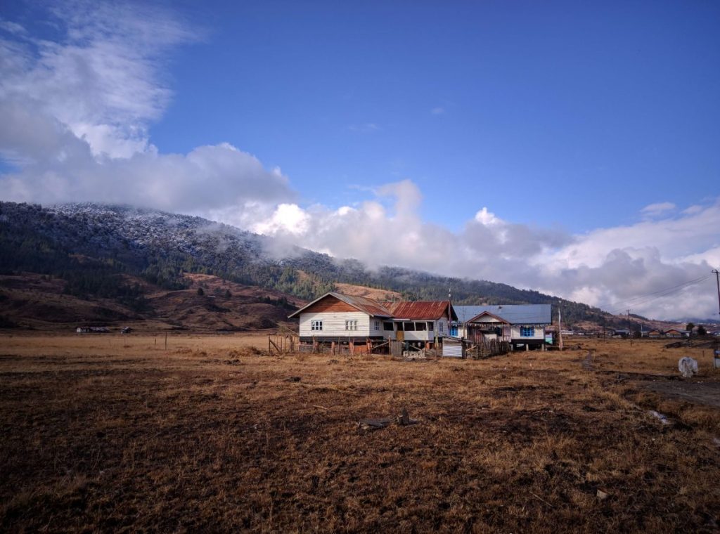 A scenic view of traditional houses in Mechuka, Arunachal Pradesh, set against a backdrop of lush green hills and clouds in a blue sky.