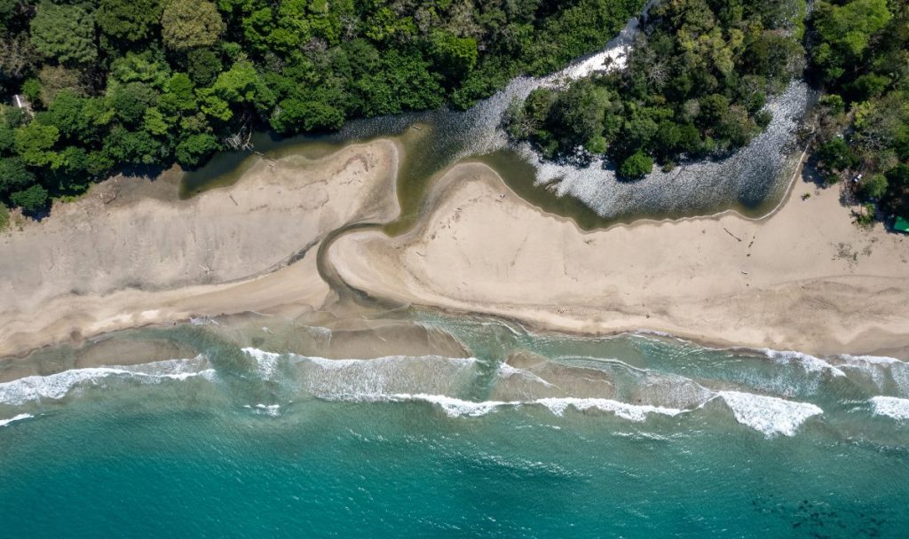 Aerial view of a sandy beach meeting the turquoise waters of the ocean, surrounded by lush green vegetation and trees.